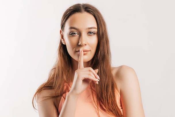 Shh gesture. Young beautiful serious girl holding a finger to her lips on a white isolated background. A woman asks to be silent, a place for advertising. Positive brunette in orange top - Fotografie, Obrázek