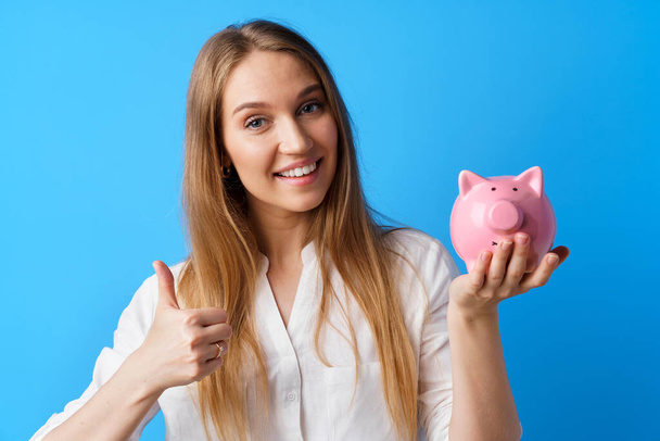 Beautiful smiling young woman with piggy bank on blue background - Photo, Image