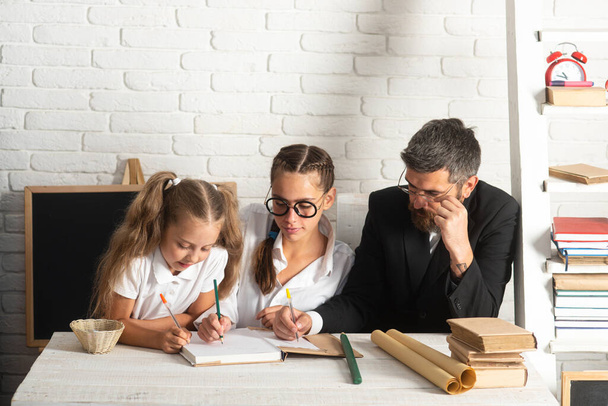 Elementary students with teacher in classroom. Father teaching schoolchild girls. Father and little kids daughters at school. Reading book and writing. - Φωτογραφία, εικόνα