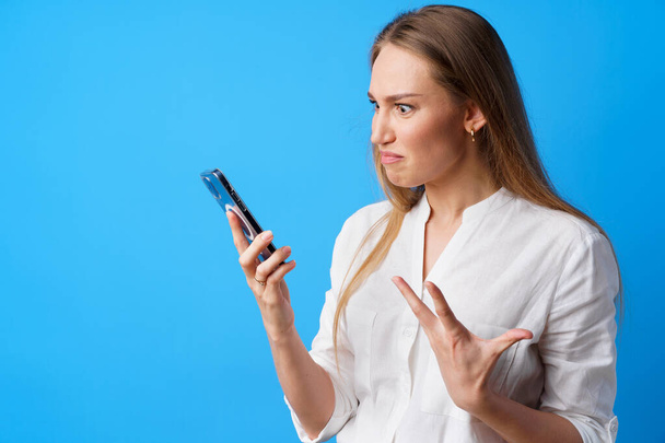 Portrait of young angry woman using her phone, annoyed texting with someone, blue background - Foto, afbeelding
