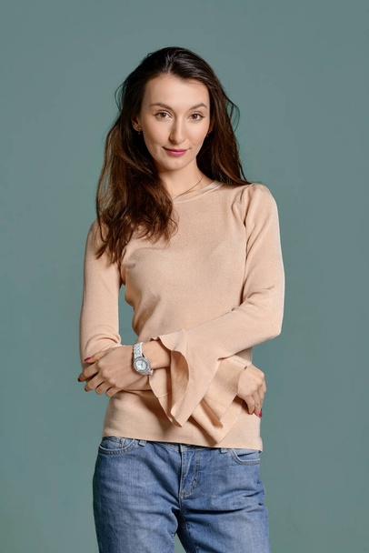 Trendy young woman in beige jersey longsleeve and straight blue jeans with crossed hands on waist - Foto, Imagem