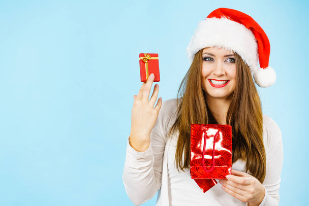 Christmas gifts. Happy woman wearing santa claus hat holding present red bag and small gift box with jewelery, on blue - Valokuva, kuva