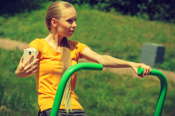 Young woman working out in outdoor gym. Girl holding mobile phone while doing exercises on street machine. - Foto, afbeelding