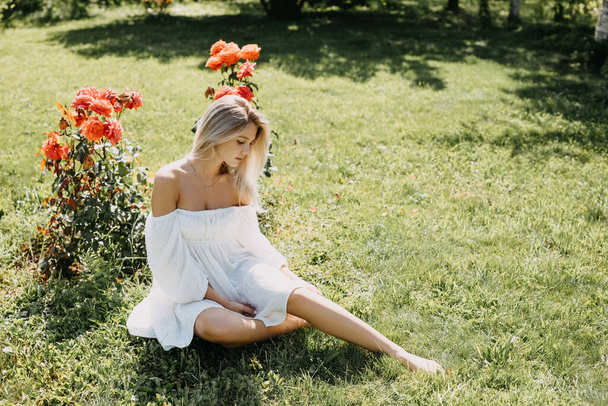 Jeune femme aux cheveux blonds assise sur l'herbe, dans un parc, par une belle journée d'été, portant une robe blanche. - Photo, image