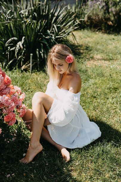 Jeune femme blonde assise sur l'herbe verte, par un jour d'été lumineux, à côté d'un buisson de roses roses, portant une robe blanche. - Photo, image