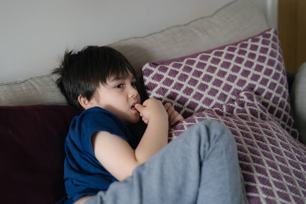 Autentický portrét Kid leží na safě a kouše si nehty, zatímco se dívá na televizi, malý chlapec leží na gauči a kouše nehty, dítě odpočívá nebo odpočívá v obýváku o víkendu - Fotografie, Obrázek