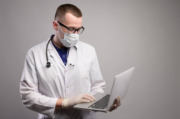 Young doctor therapist in a white coat, mask and glasses standing uses a laptop in the studio on a gray background. Portrait of a modern medic looking at the laptop screen - Photo, Image