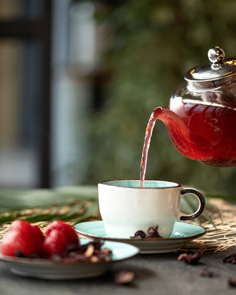 Strawberry and hibiscus natural tea. Pouring red drink from glass teapot into white clay cup. Vitamin drink for prevention of colds. Tea ceremony concept. Close up shot. Side view. Copy space - Valokuva, kuva