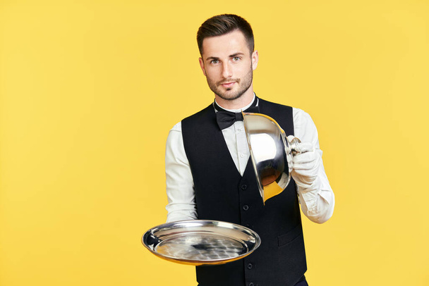 Elegant young waiter holding cloche over empty tray ready to serve on yellow background - Foto, Imagem