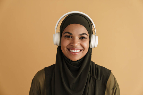 Black muslim woman in hijab listening music with headphones isolated over beige background - Фото, изображение