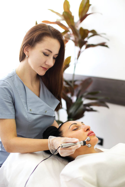 Kosmetolog stosujący nowoczesną maszynę do masażu mikroprądowego na twarzy kobiety leżącej na stole w salonie piękności - Zdjęcie, obraz