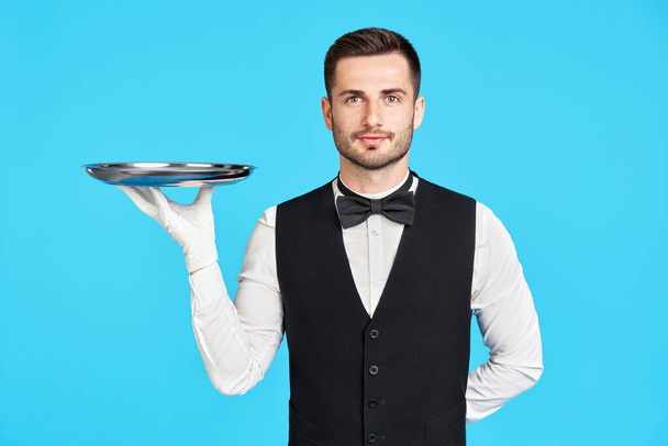 Attractive young waiter holding empty silver tray over blue background - Фото, зображення