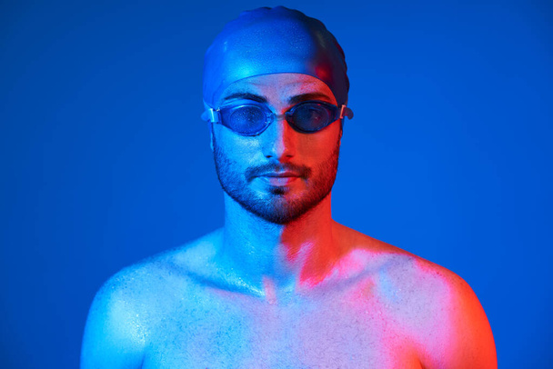 Close up portrait of handsome swimmer with goggles in red-pink neon light over blue background - Foto, Imagen