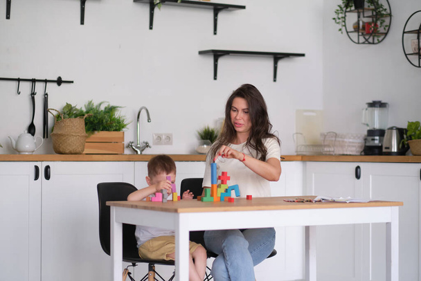 子供の男の子と母親が家のテーブルでおもちゃを遊んでいます。テーブルの上でカラフルな木製のレンガで遊ぶ子供たちは. - 写真・画像