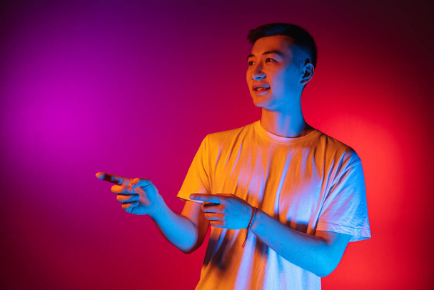 Portrait of Asian young man isolated on studio background in gradient pink purple neon light, colour filter. Concept of human emotions, facial expression. - Фото, изображение