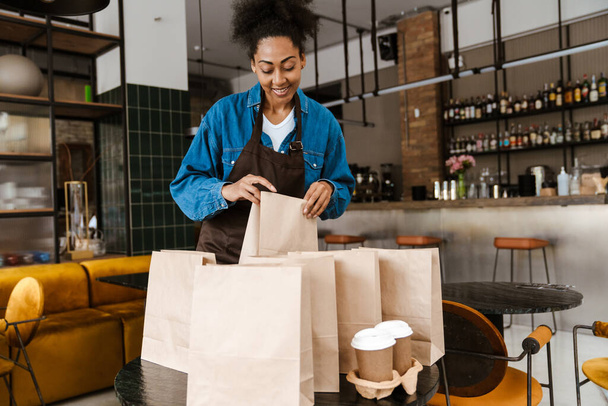Black waitress in apron packing orders while working at cafe indoors - Foto, Bild