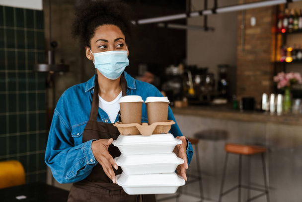 Black waitress in face mask holding food boxes while working at cafe indoors - 写真・画像
