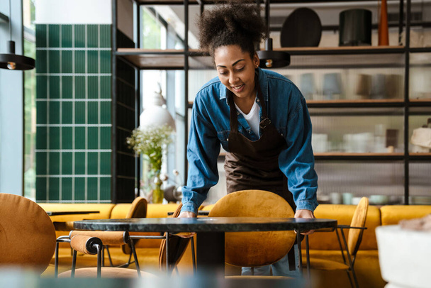 Black waitress in apron smiling while working at cafe indoors - Foto, afbeelding