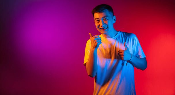 One Asian young man isolated on studio background in gradient pink purple neon light, colour filter. Concept of human emotions, facial expression. - 写真・画像