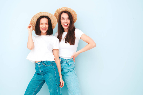 Two young beautiful smiling hipster female in trendy summer white t-shirt and jeans clothes.Sexy carefree women posing near light blue wall in studio.Positive models in hats. Happy and cheerful - Φωτογραφία, εικόνα
