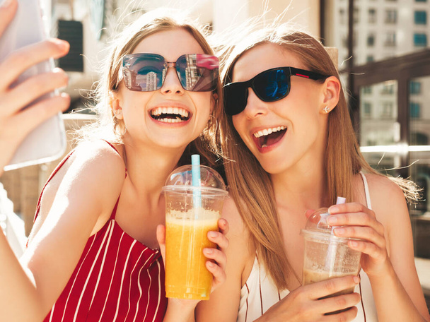 Two young beautiful smiling hipster female in trendy summer clothes. Carefree women posing outdoors.Positive models drinking fresh cocktail smoothie drink in plastic cup with straw.Taking selfie - Photo, image
