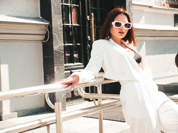 Portrait of young sexy modern businesswoman model. Hot beautiful woman in white suit posing on the street background. Fashion female walking outdoors in sunglasses - Фото, изображение