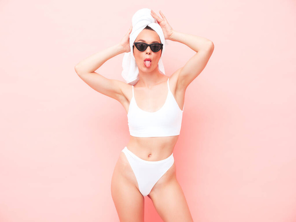 Young beautiful smiling woman in white lingerie. Sexy carefree model in underwear and towel on head posing pink wall in studio. Positive and happy female enjoying morning in sunglasses.Shows tongue - Φωτογραφία, εικόνα