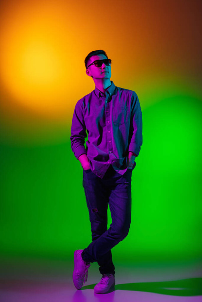 Young man, fashion model in casual clothes isolated on studio background in gradient green yellow neon light, colour filter. Concept of human emotions, facial expression. - Фото, изображение