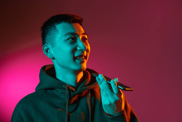Полудлинный портрет азиатского юноши, изолированный на студийном фоне в градиентном красном неоновом свете, цветной фильтр. Концепция человеческих эмоций, выражение лица. - Фото, изображение