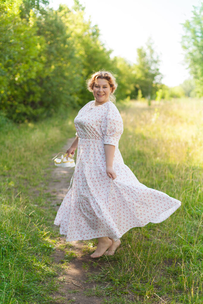 a young happy woman walks barefoot in a field on a summer day, holding shoes in her hands. Model plus size. selective focus. - Photo, Image