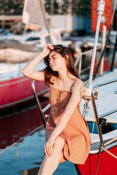 νεαρή κοπέλα με φόρεμα κάθεται σε μια βάρκα σε μια προβλήτα στη θάλασσα το βράδυ του καλοκαιριού, ο ήλιος που δύει, διακοπές - Φωτογραφία, εικόνα