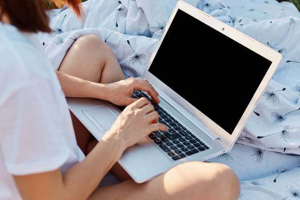 Faceless unknown woman wearing white casual t shirt sitting with crossed legs and holding laptop with black blank screen, copyspace for advertisement, female freelancer working outdoor. - 写真・画像