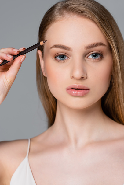 young woman applying brown eyebrow pencil isolated on gray - 写真・画像
