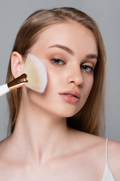 fiatal nő gazdaság kozmetikai kefe és alkalmazása arc por elszigetelt szürke - Fotó, kép