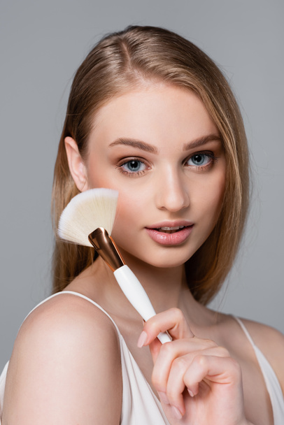 fiatal és csinos nő gazdaság kozmetikai kefe elszigetelt szürke - Fotó, kép