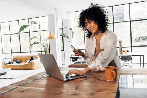 Fiatal, gyönyörű afro-amerikai nő gépel laptopon a konyhapulton, kávézik és tartja a mobilját. Másold a helyet. Otthon koncepció. Technológiai koncepció. - Fotó, kép