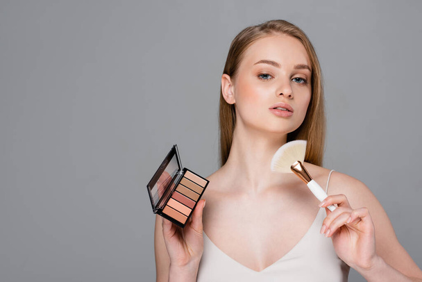 pretty woman holding cosmetic brush and eye shadow and blush palette isolated on grey - Valokuva, kuva