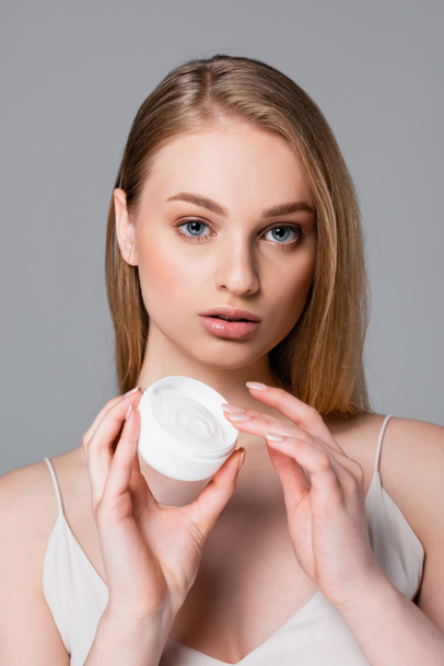 pretty young woman holding container with cosmetic cream isolated on gray - Photo, Image