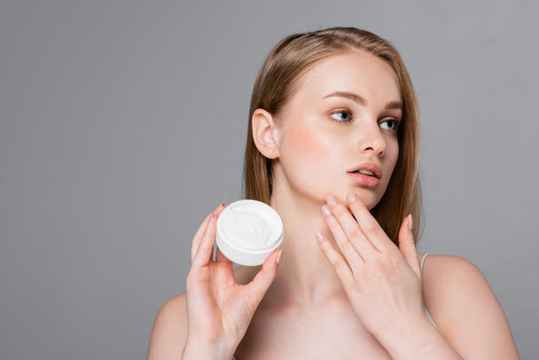 pretty young woman holding container with cosmetic cream and looking away isolated on grey - Photo, Image