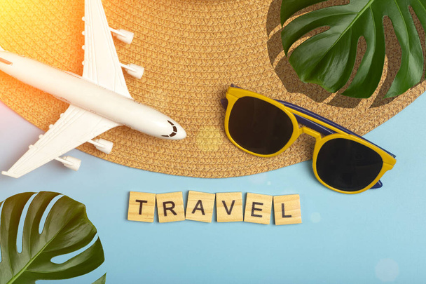 Ταξίδι καλοκαιρινό φόντο. Γυαλιά ηλίου, καπέλο, παλάμη τροπικά φύλλα, αεροπλάνο και αξεσουάρ παραλία σε ένα χρωματιστό μπλε φόντο. Τουριστικές διακοπές, χαλάρωση και καλοκαιρινή έννοια. - Φωτογραφία, εικόνα