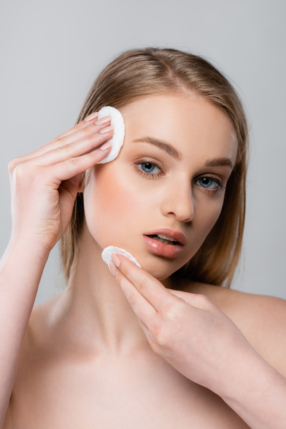 young woman with bare shoulders removing makeup with cotton pads isolated on gray - Foto, Bild