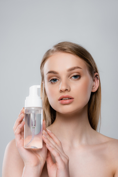 young woman with bare shoulders holding bottle with micellar water isolated on grey - Foto, Imagen