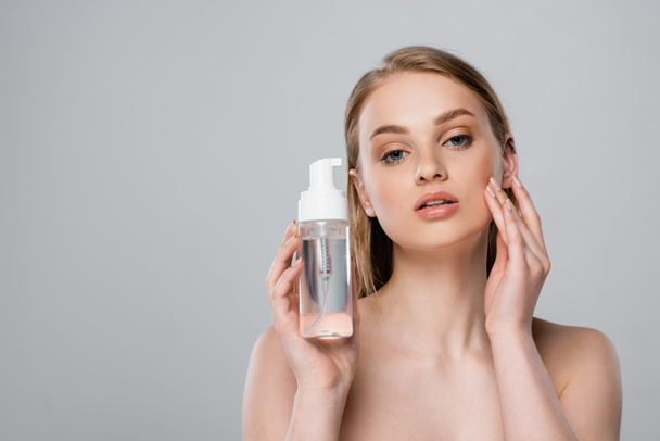 pretty young woman with bare shoulders holding bottle with micellar water isolated on grey - Zdjęcie, obraz