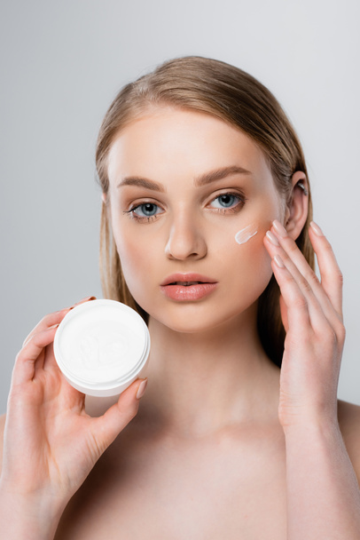 pretty woman with blue eyes applying cream on face isolated on grey - Photo, Image