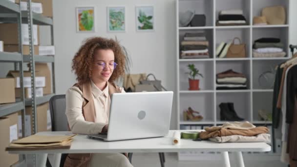 malý byznys, portrét mladé ženy přijímající objednávky online na notebooku, zatímco sedí u stolu v obchodě s oblečením - Záběry, video