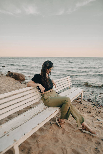 Спокойная девушка с темными волосами сидит на белой скамейке на берегу моря. - Фото, изображение