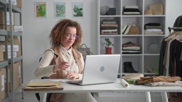 portrét půvabné mladé ženy přijímající objednávky z internetového obchodu, zatímco sedí u notebooku v modg butiku, usmívá se a dívá se do kamery - Záběry, video