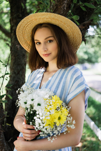 Jeune femme en chapeau de soleil tenant des fleurs dans le parc  - Photo, image