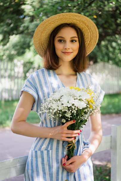 Femme souriante en chapeau de soleil et robe tenant bouquet dans le parc  - Photo, image