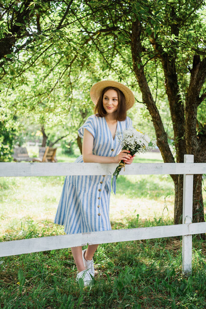 Smiling woman in sun hat holding bouquet near fence in park  - Photo, Image
